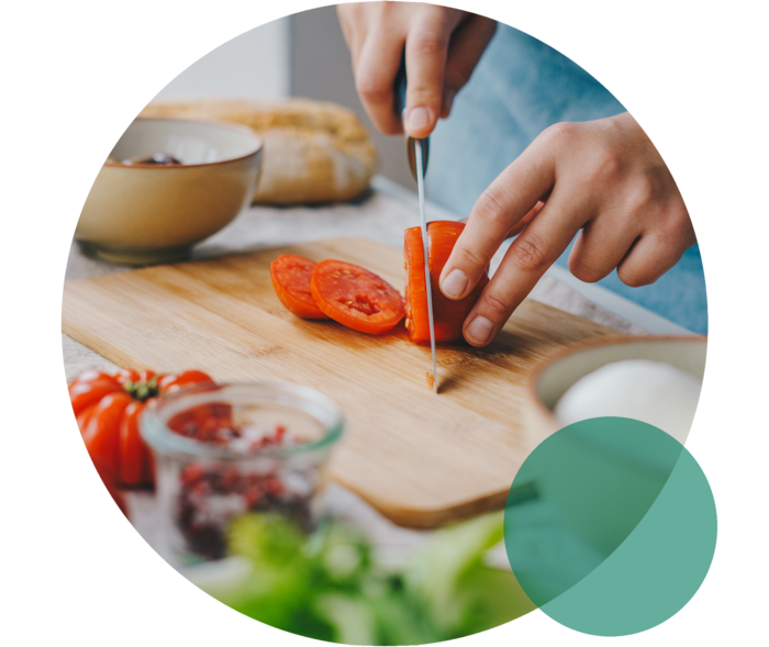 Ernährung bei Hypercholesterinämie Küchenbrett auf einer Arbeitsplatte. Zwei Hände schneiden eine Tomate mit einem Messer.