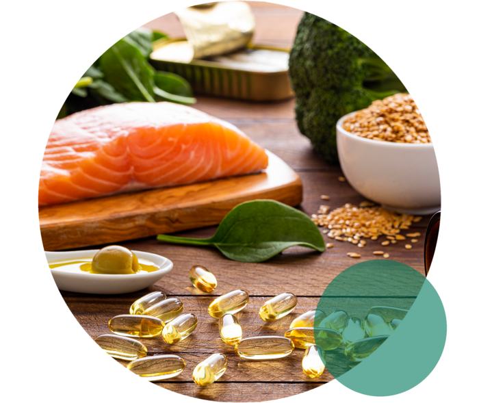 Erhöhte Triglyceride: Gesunde Ernährung mit Omega-3-Fettsäuren Auf einer Fläche liegen Lachs, Blattspinat, Brokkoli, ein Löffel mit Olivenöl sowie gelbliche Kapseln.