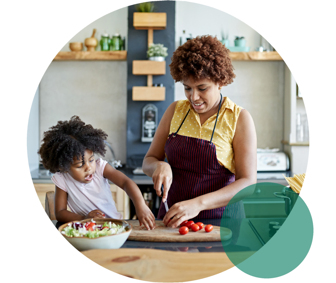 Hohes Cholesterin: Die Rolle einer ausgewogenen Ernährung Eine Frau mittleren Alters mit Küchenschürze. Sie schneidet lächelnd Tomaten. Neben ihr steht ein Kind, das ihr zusieht.
