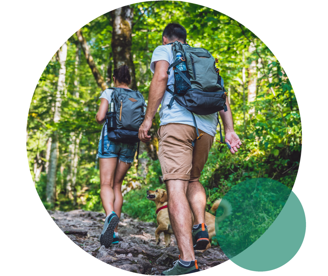 Einfluss der Bewegung auf das Cholesterin Eine junge Frau und ein junger Mann in sommerlicher Wanderbekleidung. Sie gehen bergauf auf einem Wanderweg, der von Bäumen umsäumt ist.