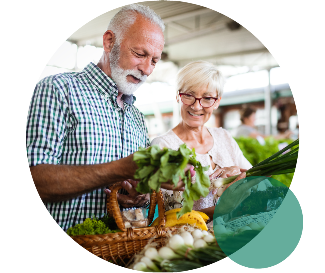 Sekundäre Hypercholesterinämie: Die Bedeutung des Lebensstils Ein älteres Ehepaar steht lächelnd an einem Marktstand mit Gemüse. Der Mann hält einen Salatkopf in der Hand, den beide betrachten.