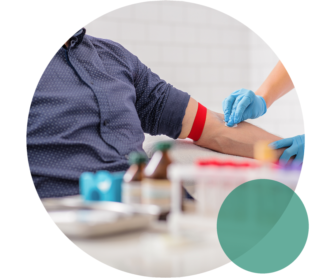 Blutabnahme zur Diagnose erhöhter Cholesterinwerte Ein Mann sitzt auf einem Stuhl. Neben ihm steht eine Person in weißem Kittel. Sie trägt Einweghandschuhe und streift mit einem Alkoholtupfer über die linke Armbeuge des Manns.